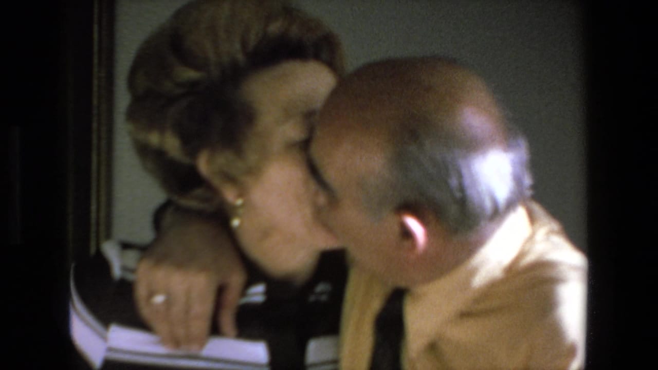 Elderly couple kissing while seated closely together inside a warmly lit room.