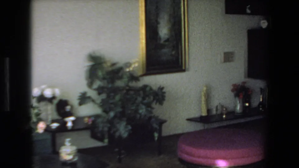 1972 Mazatlan home-movie frame showing a decorated room with a large plant, framed wall art, small display shelf, dark cabinet, and pink circular ottoman.