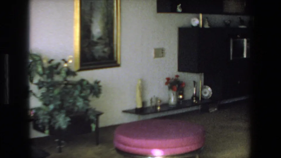 1972 Mazatlan home-movie frame of a decorated interior corner with a prominent pink round ottoman, leafy plant, framed painting, display shelf, and dark cabinet.