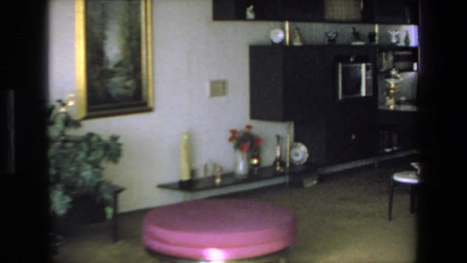 1972 Mazatlan home-movie frame showing a furnished living room with a dark wall unit, framed painting, potted plant, low shelf, chair, and pink round ottoman.