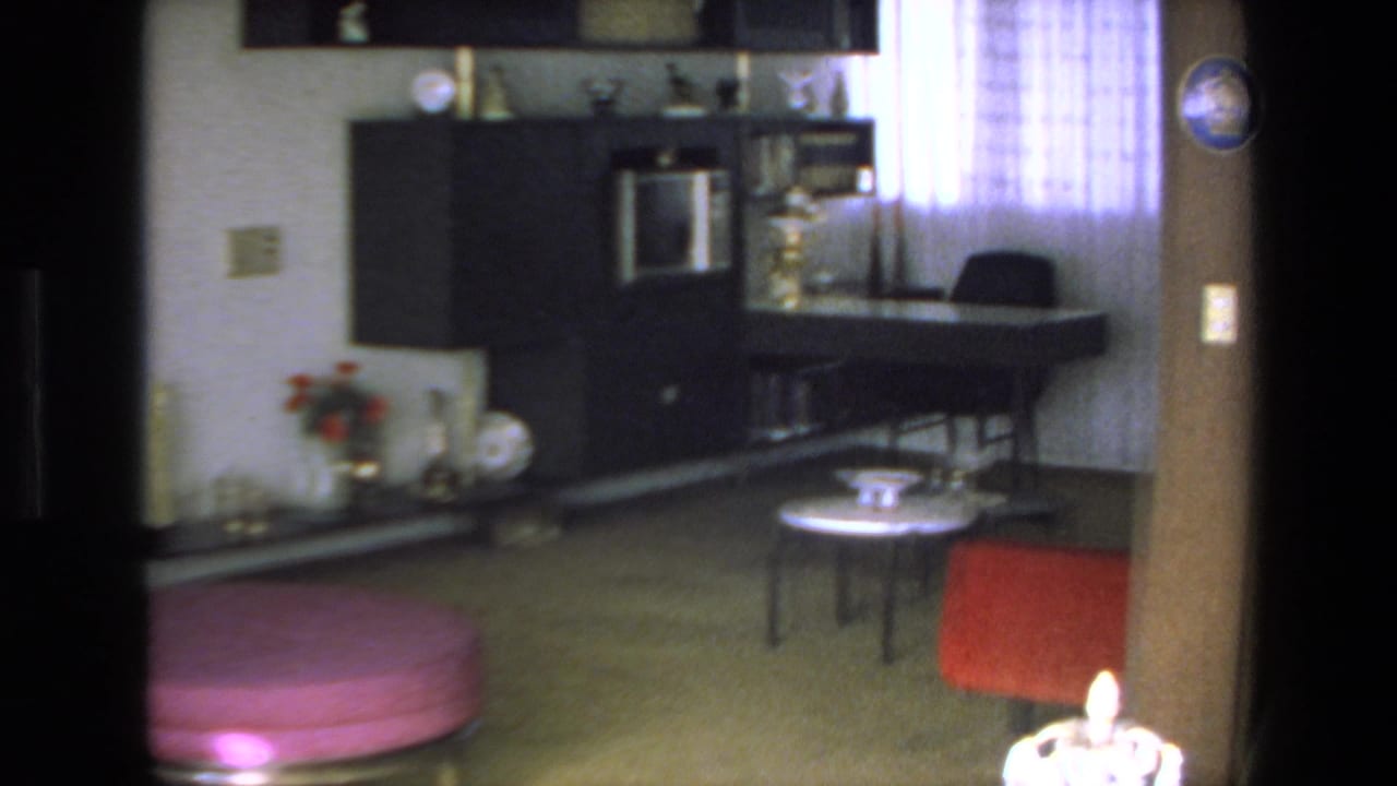 1972 Mazatlan interior frame with a dark wall unit, desk or counter area by a sheer curtained window, small round table, and red upholstered chair.