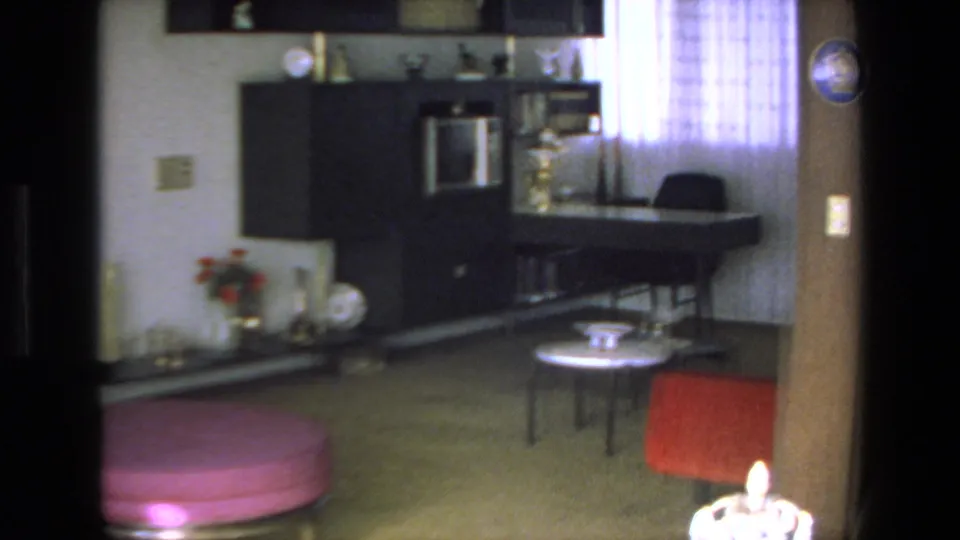 1972 Mazatlan interior frame with a dark wall unit, desk or counter area by a sheer curtained window, small round table, and red upholstered chair.