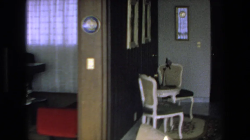 1972 Mazatlan home interior showing a curtained window, red chair, dark paneled doors, two white chairs with dark cushions, wall art, and a small table or rug area.