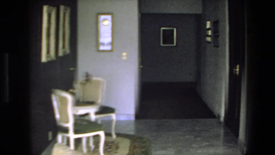 1972 Mazatlan interior frame looking toward a hallway with two white chairs, framed wall decor, a dark doorway, light-colored floor, and a corridor receding into the home.