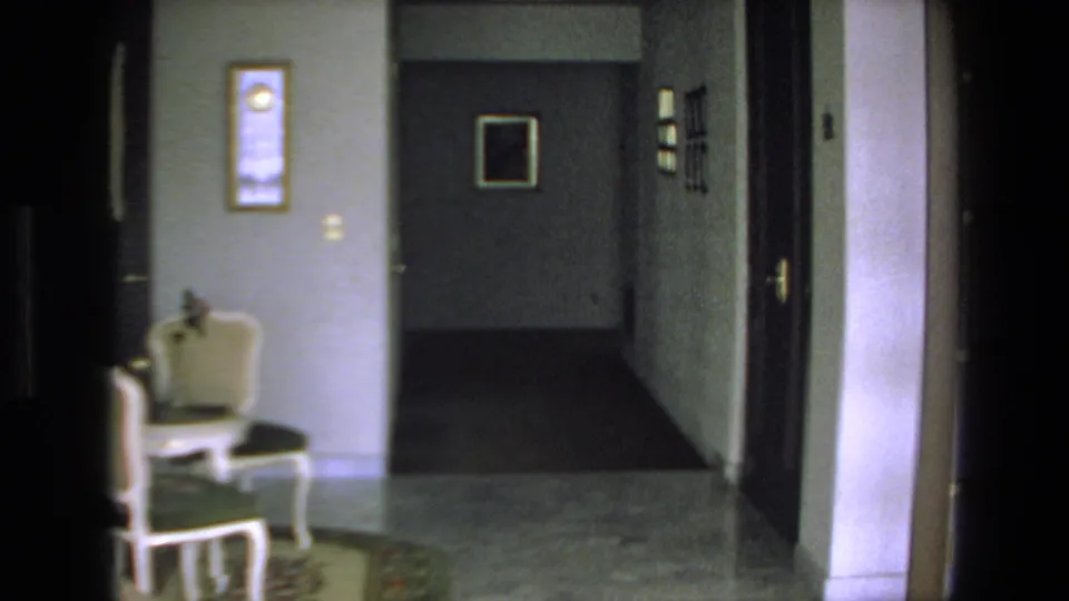 A dim 1972 Mazatlan home interior shows a tiled hallway, white upholstered chair, framed wall art, and several doorways in soft 8mm focus.