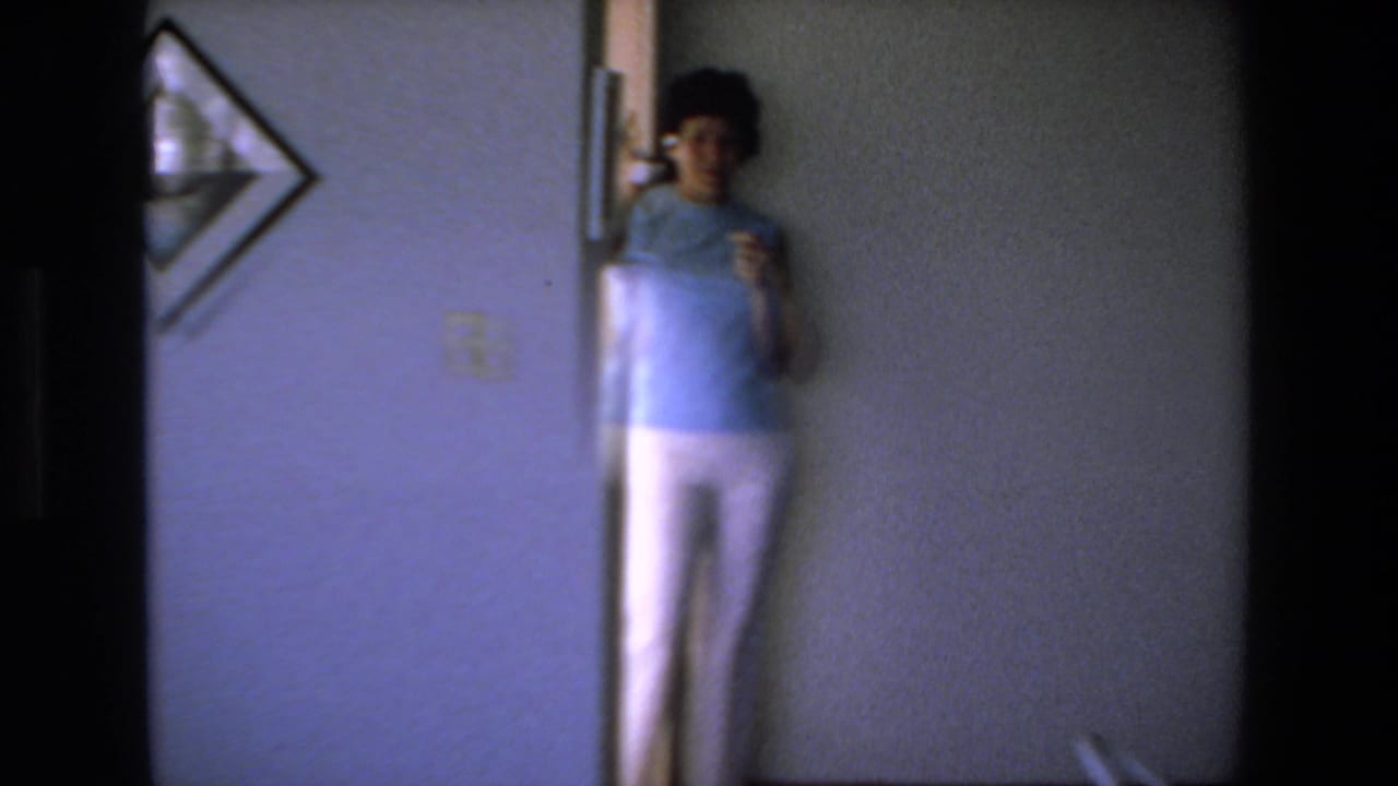 Woman standing near a doorway and wall telephone in a dim interior.