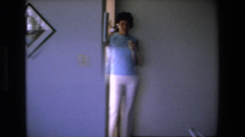 A woman in a light blue top and white pants stands near a doorway and wall telephone inside the Mazatlan home interior.