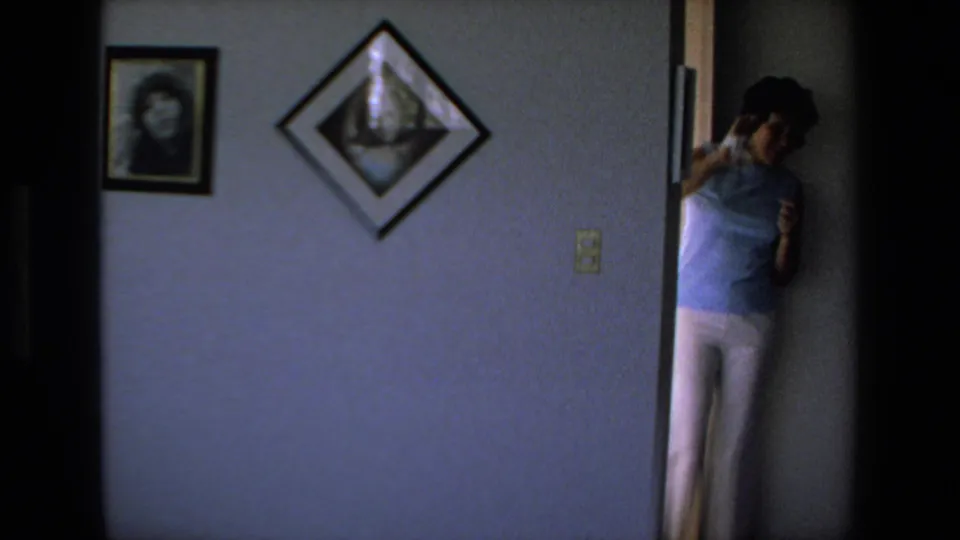 The woman appears partly in a doorway beside framed pictures and diamond-shaped wall art in the softly lit 1972 home interior.