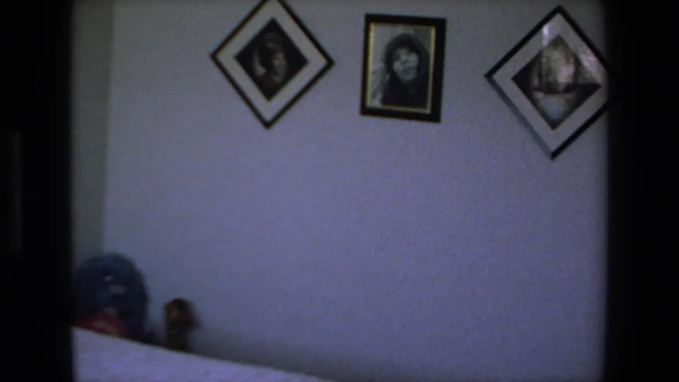 1972 Mazatlan home-movie frame showing a dim bedroom wall with framed pictures, part of a bed, and a soft object near the head of the bed.