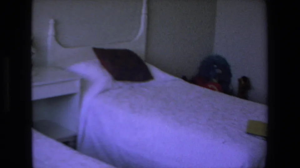 1972 Mazatlan home-movie frame of a twin bed with a white headboard, dark pillow, pale bedding, and bedside table in low light.