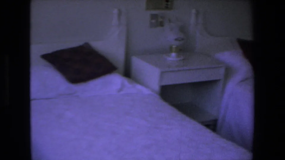 1972 Mazatlan home-movie frame showing twin beds with pale bedding, a white nightstand between them, and a small bedside lamp or object.