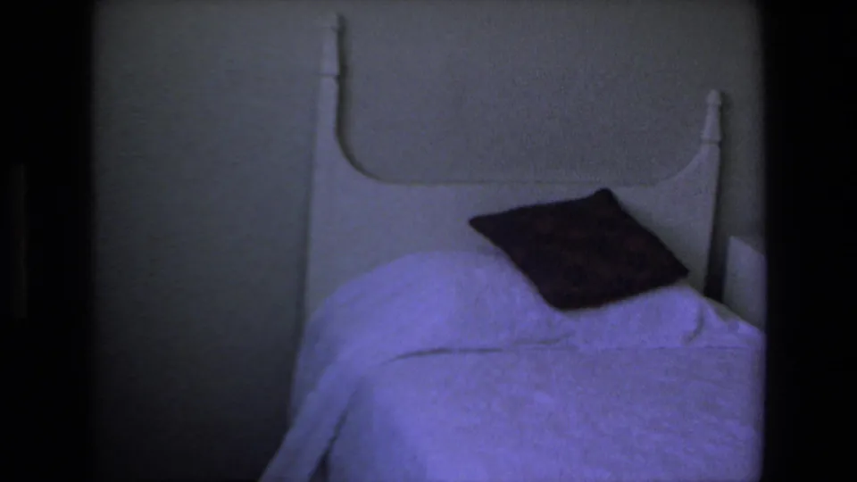 1972 Mazatlan home-movie frame focused on a twin bed with a white headboard, pale bedspread, and dark pillow in a dim private room.