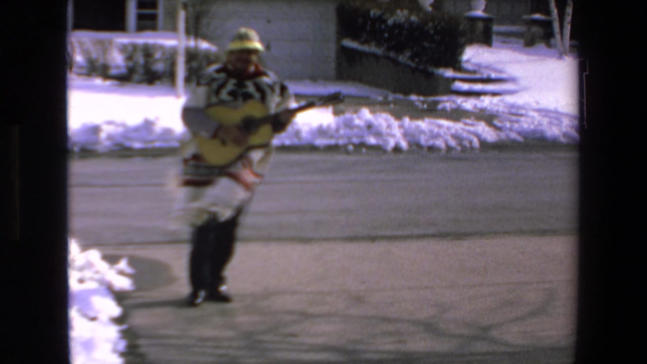 Man in a patterned poncho and hat walks on a snowy street carrying a guitar.
