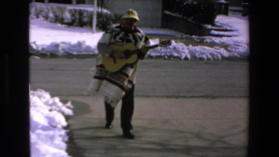 The man in a patterned poncho appears to strum a guitar while walking past snowbanks on a residential street in the 1972 Mazatlan clip.