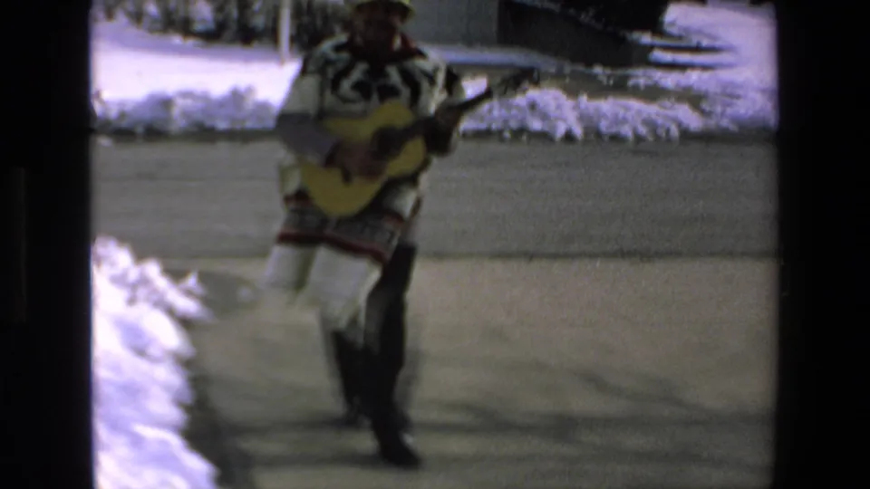 The man with the guitar moves closer in soft 8mm focus, with snow piled along the edge of the residential lane behind him.