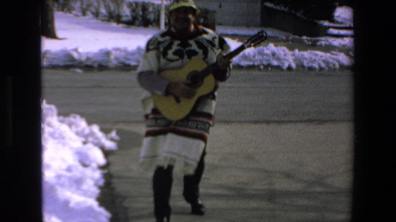 In 1972 Mazatlan home-movie footage, a man in a patterned poncho and yellow hat walks on a snow-lined driveway while holding and playing an acoustic guitar.