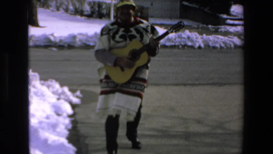 In 1972 Mazatlan home-movie footage, a man in a patterned poncho and yellow hat walks on a snow-lined driveway while holding and playing an acoustic guitar.