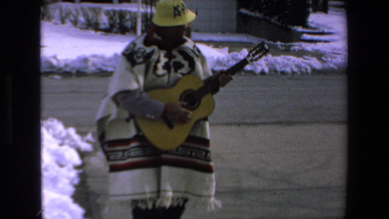 Man in a poncho and yellow hat stands with an acoustic guitar near snowbanks and pavement.
