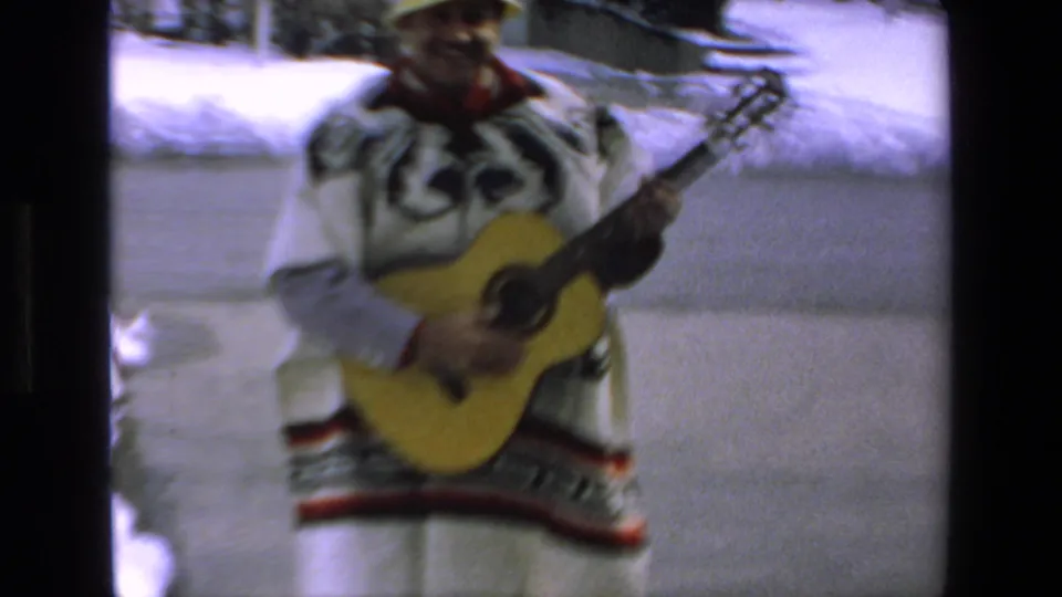 The 1972 Mazatlan footage moves closer to the man as he plays an acoustic guitar, with his patterned poncho, yellow hat, snow, and driveway filling the frame.