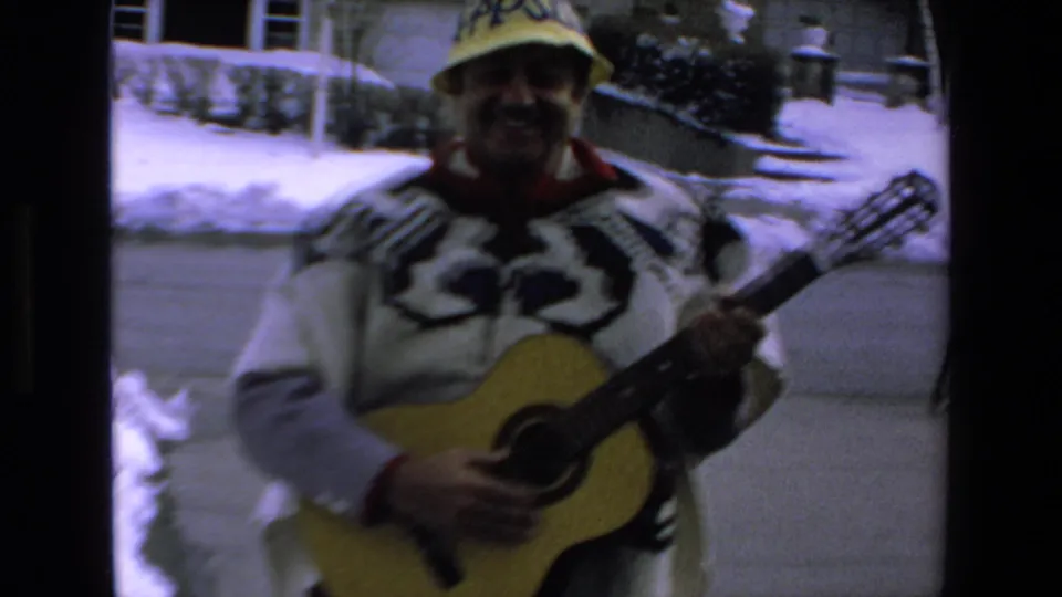 A close 1972 Mazatlan home-movie frame shows the smiling man in a yellow hat and patterned poncho holding his acoustic guitar near snow-covered pavement.