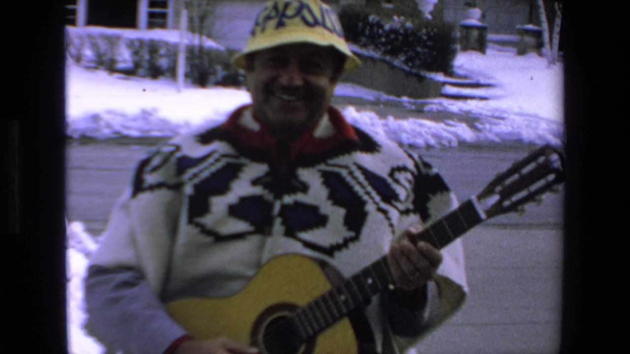 In supplied 1972 Mazatlan home-movie footage, a smiling adult man wearing a patterned sweater and yellow hat holds an acoustic guitar outside near snow and a house exterior.