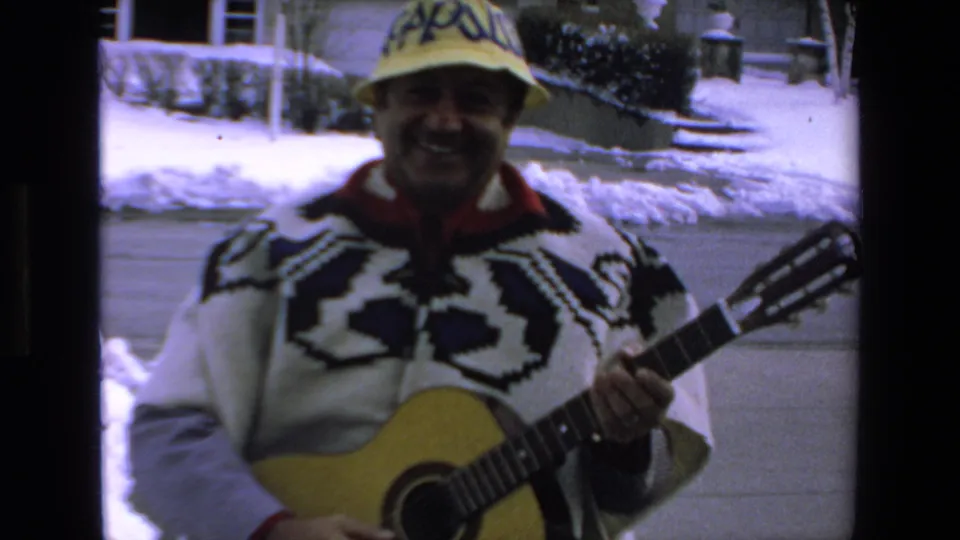 In supplied 1972 Mazatlan home-movie footage, a smiling adult man wearing a patterned sweater and yellow hat holds an acoustic guitar outside near snow and a house exterior.