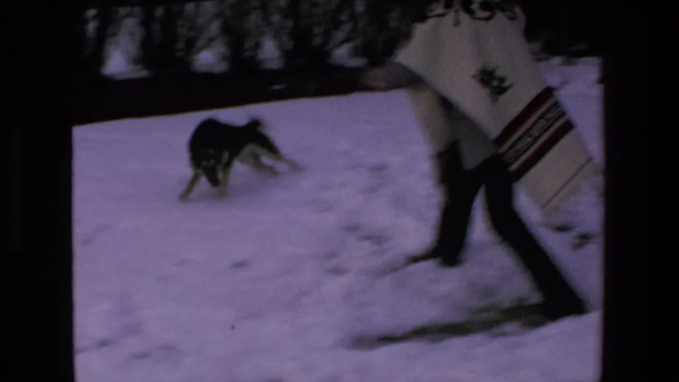 A person wearing a patterned poncho walks across the snowy yard while the dog turns nearby in the 8mm home-movie frame.
