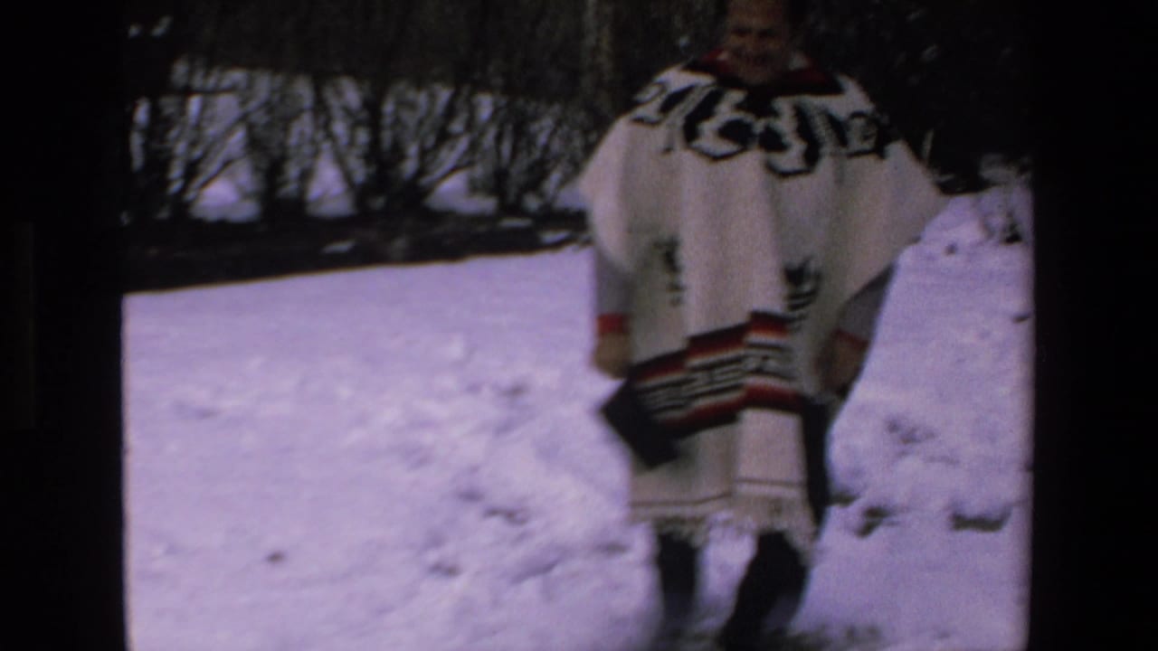 Man in a patterned white poncho standing on snow with dark trees behind him.