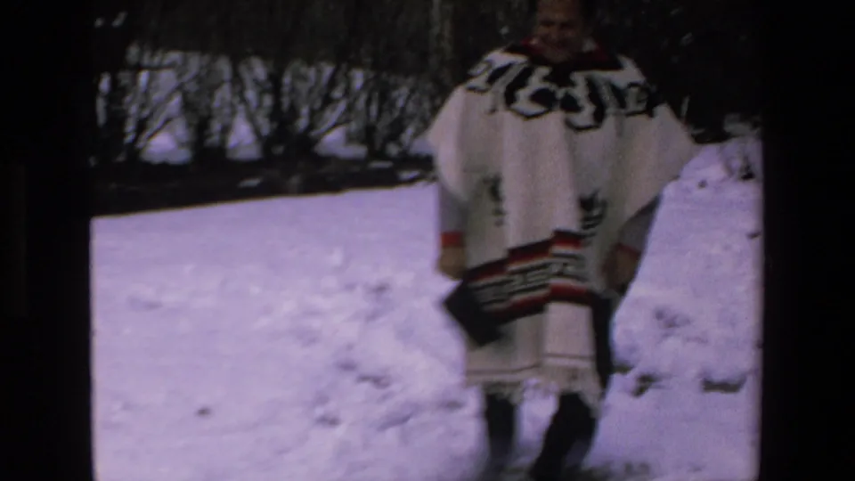 A man in a white poncho with black and red geometric trim stands on snow in the supplied 1972 Mazatlan home-movie footage.
