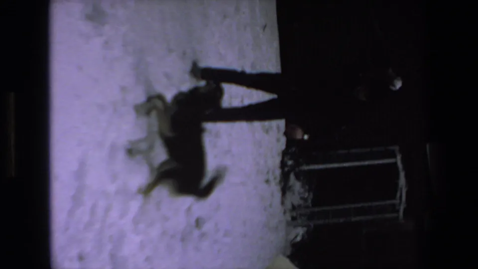 The dog moves across the snowy ground as an adult stands nearby, recorded in soft, grainy 8mm low-light footage.