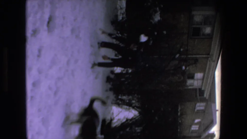 In this 1972 8mm home-movie frame, a dark dog appears mid-leap across a snowy residential yard with shrubs and part of a house visible behind it.