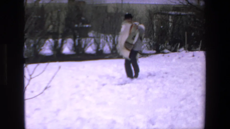 A person wearing a patterned poncho stands in the snow in a residential yard, with bare bushes and fencing behind them in the supplied 1972 home-movie context.