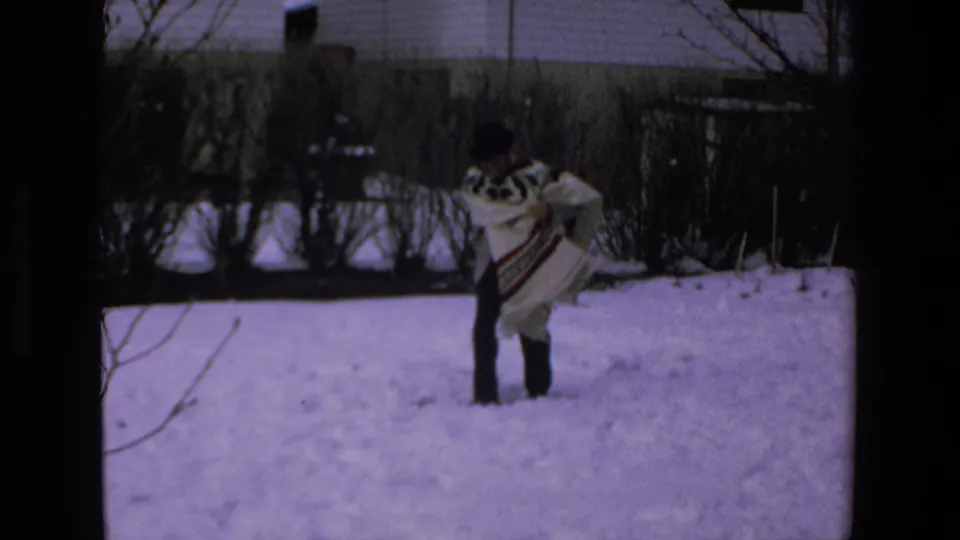 The person in the patterned poncho remains in the snowy yard, turned slightly near bushes and fencing, with the soft grain and color cast of 8mm film visible.