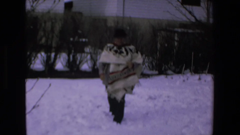 Adult man wearing a white patterned poncho and dark hat is seen from behind while walking across a snowy residential yard in the supplied 1972 footage.