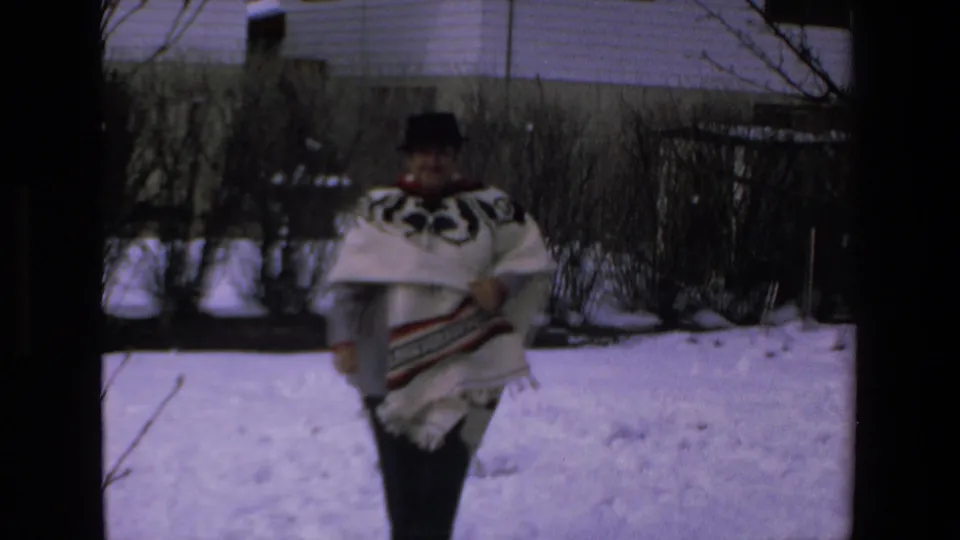 Adult man in a white geometric-patterned poncho and dark hat approaches the camera through snow, with bare bushes and a house behind him.