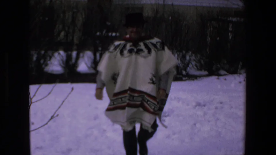 Adult man in a white patterned poncho and dark hat fills more of the frame as he moves through the snow in the 8mm home-movie scene.