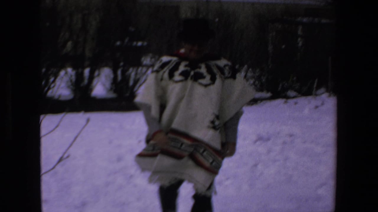 In 1972 8mm footage identified as Mazatlan, Mexico, an adult man in a white poncho with black geometric patterns moves through a snow-covered yard with bare shrubs behind him.
