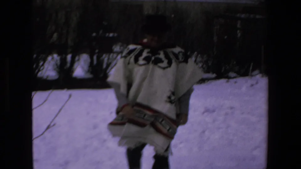 In 1972 8mm footage identified as Mazatlan, Mexico, an adult man in a white poncho with black geometric patterns moves through a snow-covered yard with bare shrubs behind him.