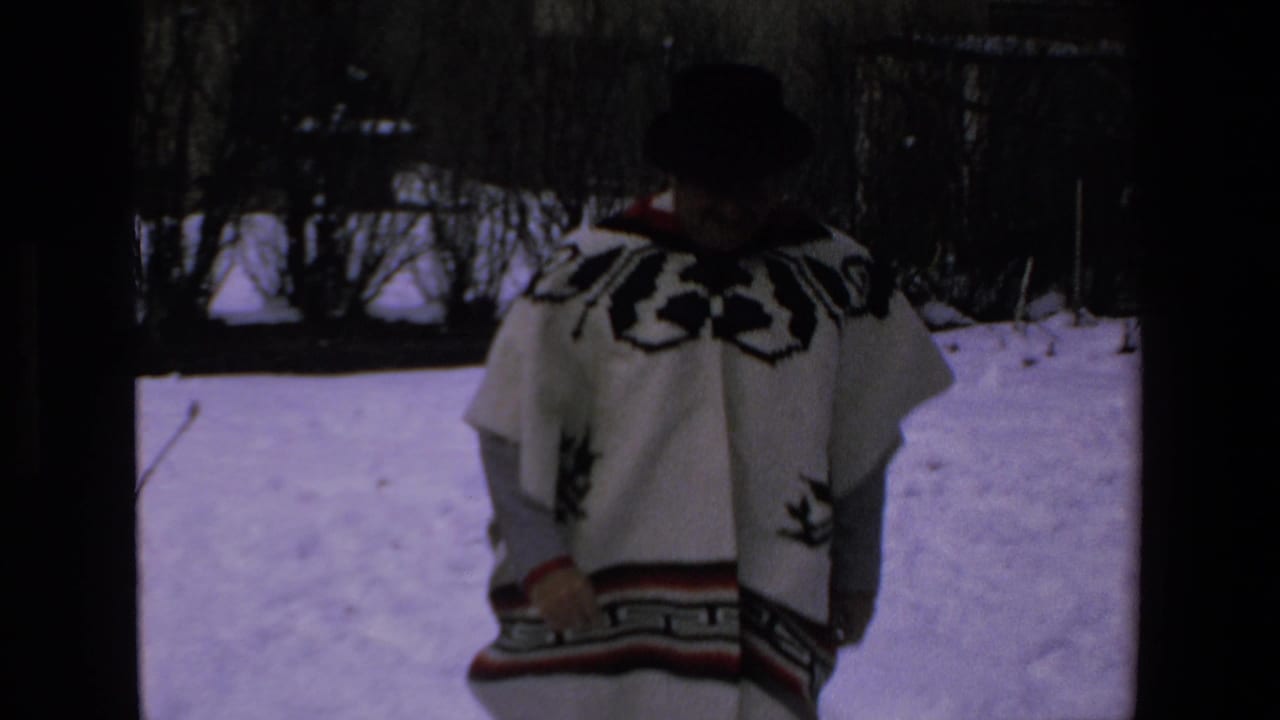Man in a white patterned poncho stands in profile in snow.