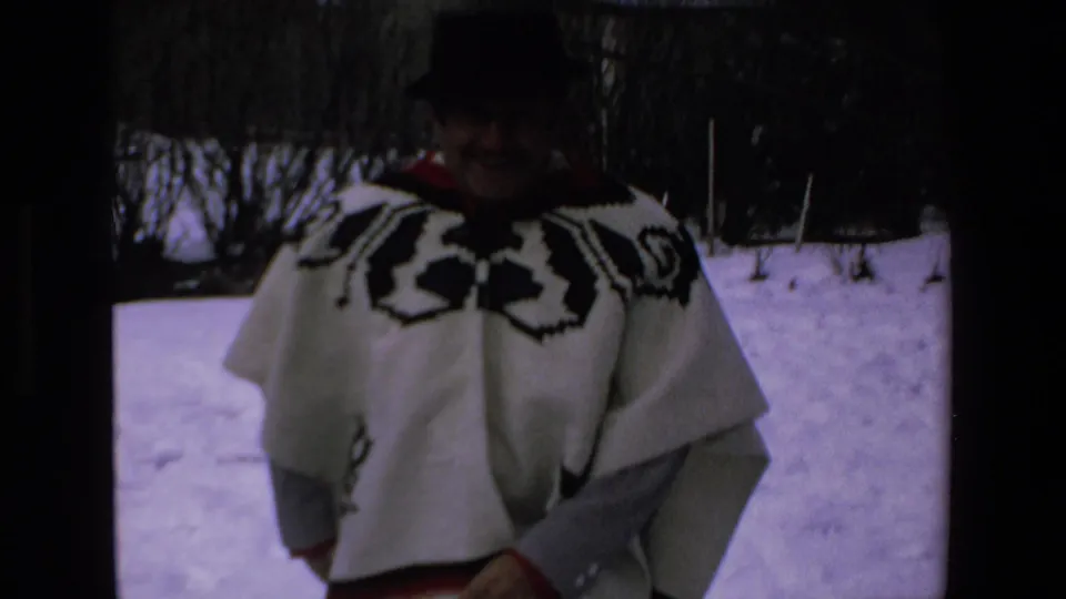 The man faces the handheld camera while wearing a white poncho with black geometric designs and a dark hat, with snow and bare winter shrubs visible behind him.