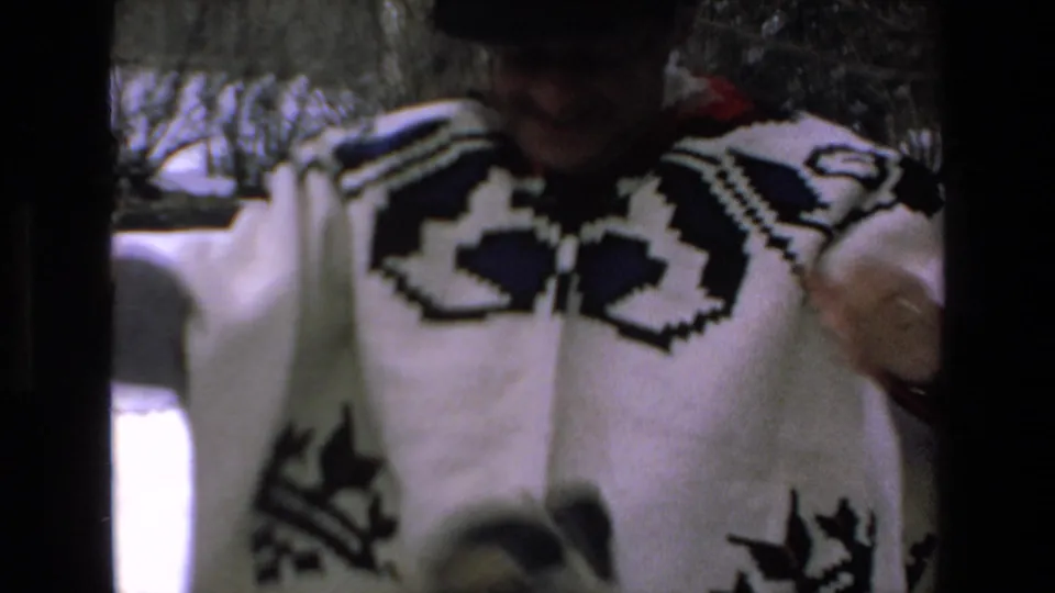 In 1972 8mm home-movie footage identified as Mazatlan, Mexico, a man in a black hat and bold patterned sweater stands in snow near bare trees, partly shadowed by the frame.