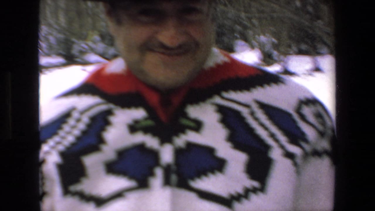 Smiling man in a black hat and patterned sweater stands close to the camera in snow.