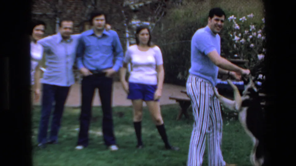 The man turns with the stick while the black-and-white dog moves beside him, with adult friends still visible in the 1972 Mazatlan garden scene.