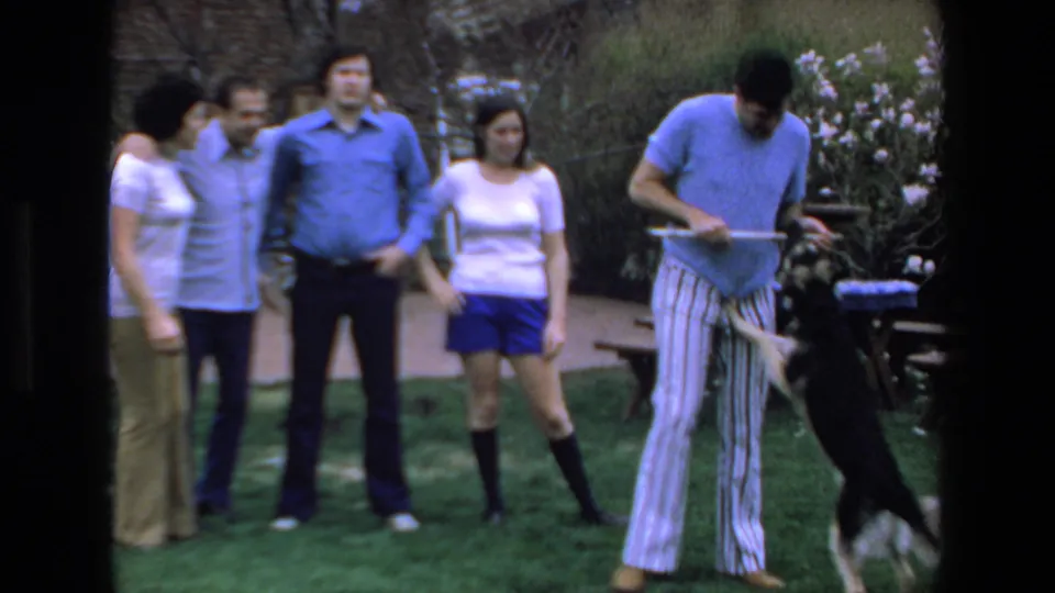 In 1972 Mazatlan home-movie footage, adults in casual clothing stand on a grassy lawn as a man in a blue shirt and striped pants plays with a black-and-white dog.