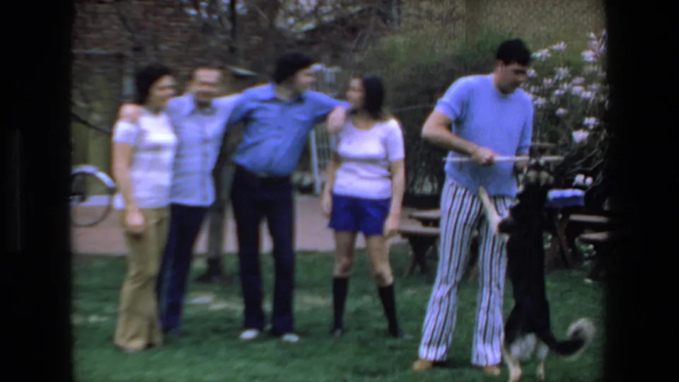 The 1972 Mazatlan home-movie frame shows adults gathered casually while a man continues playing with a black-and-white dog on a lawn near picnic tables.