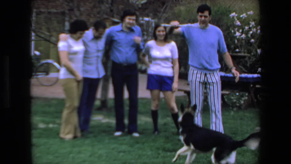 In documented 1972 Mazatlan home-movie footage, five casually dressed adults stand in a backyard as a German Shepherd-type dog runs across the grass.