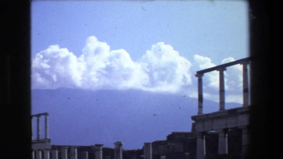 1973 Rome 8mm frame showing ancient masonry and partial classical columns viewed through a dark window or doorway edge, with distant hills and bright white clouds behind.