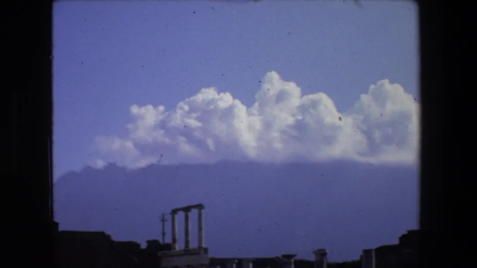 1973 Rome 8mm view of low ancient ruins and column fragments beneath a blue sky, with hazy hills and clustered white clouds in the background.