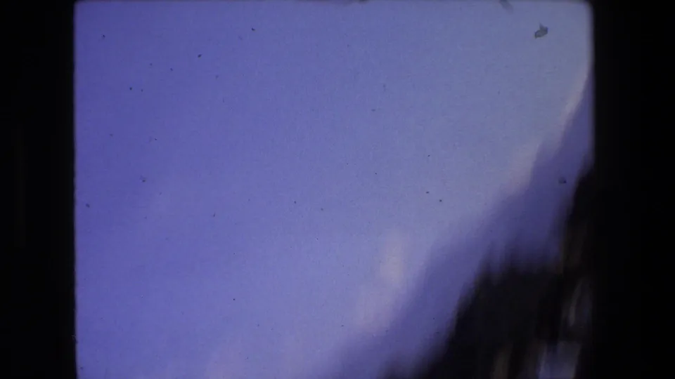 Soft, partially obscured 8mm frame from the Rome ruin sequence, showing a pale blue field, dark curved edge, and visible film specks rather than a clear subject.