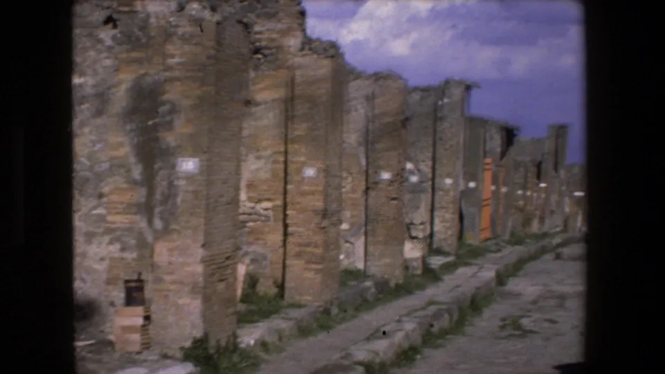 1973 Rome 8mm frame showing a long weathered ancient wall beside a narrow path, with rough masonry, small openings, vegetation, and blue sky visible above.